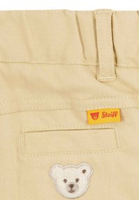 Pantaloni in cotone beige con tasca cucita, patch a forma di testa d'orso bianco e etichetta gialla con logo dell'orso Steiff.