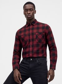 Man draagt een rood-zwart geruit overhemd met lange mouwen en zwarte jeans, staand tegen een effen lichte achtergrond.