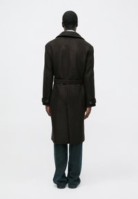 Abrigo de lana marrón oscuro con cuello, cintura ceñida con cinturón y puños abotonados, visto desde atrás. Combinado con pantalones de mezclilla de pierna ancha.