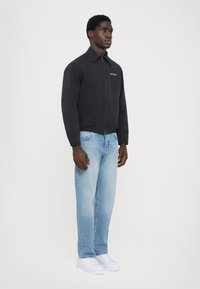 Black zip-up jacket with a collar, paired with light blue straight-leg jeans and white sneakers; minimal stitching and logo detail.