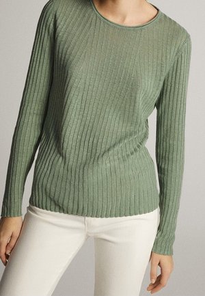 Femme portant un pull ajusté à manches longues en maille côtelée vert olive et un pantalon écru, debout devant un fond neutre.