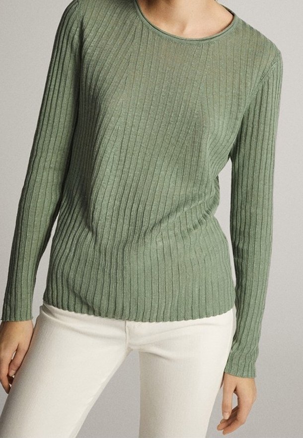 Femme portant un pull ajusté à manches longues en maille côtelée vert olive et un pantalon écru, debout devant un fond neutre.