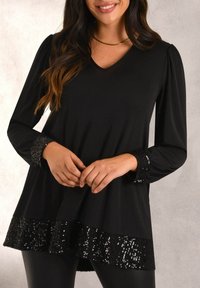 Black long-sleeve top with a V-neckline, featuring textured sequin accents at the sleeves and hem, with a smooth, flowing fabric.