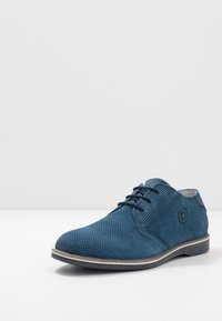 Chaussure en cuir bleu perforée avec un bout arrondi, des lacets noirs et une semelle blanche. Elle présente un petit logo sur le côté.