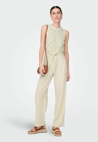 Top sin mangas de punto verde claro con lazo en el frente, combinado con pantalones anchos beige, bolso cruzado marrón y sandalias de plataforma marrones.