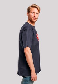 Navy blue short-sleeve t-shirt featuring a red graphic design, with a relaxed fit and classic round neckline. Worn with light denim jeans.