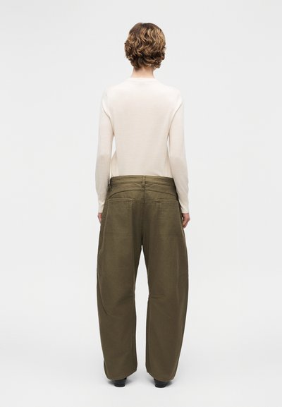 Άτομο με κοντά σγουρά μαλλιά, φοράει μακρυμάνικη μπεζ μπλούζα και φαρδιά χακί παντελόνια, στέκεται γυρισμένο πλάτη σε απλό φόντο.