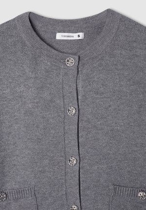 Szary sweter z dzianiny, z okrągłym dekoltem, pięcioma dekoracyjnymi guzikami i dwiema przednimi kieszeniami. Posiada ściągacze przy mankietach i na dole. Widoczna etykieta marki.