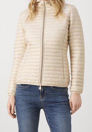 Femme portant une veste matelassée beige zippée et un jean bleu, affichée des épaules aux hanches sur un fond uni.