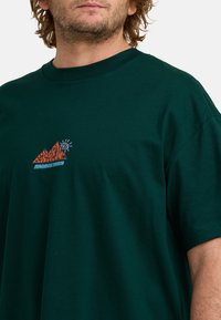 Camiseta de algodón verde oscuro con cuello redondo, que presenta un diseño gráfico colorido de montañas y un ojo en el pecho.