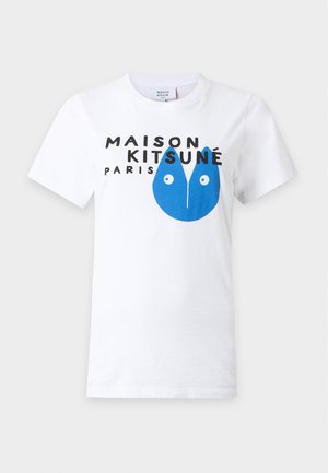 Femme portant des lunettes de soleil noires, un t-shirt blanc à motif graphique, un pantalon beige à jambes larges, tenant un sac cabas noir sur un fond clair uni.