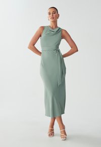 Long, flowy dress in muted green fabric with a draped neckline, fitted waist, and side tie detailing. Paired with white strappy heels.