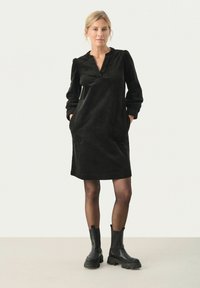 Robe en velours côtelé noire avec un décolleté en V, des manches bouffantes et des poches latérales. Associée à des bottines noires. Texture lisse et coupe décontractée.