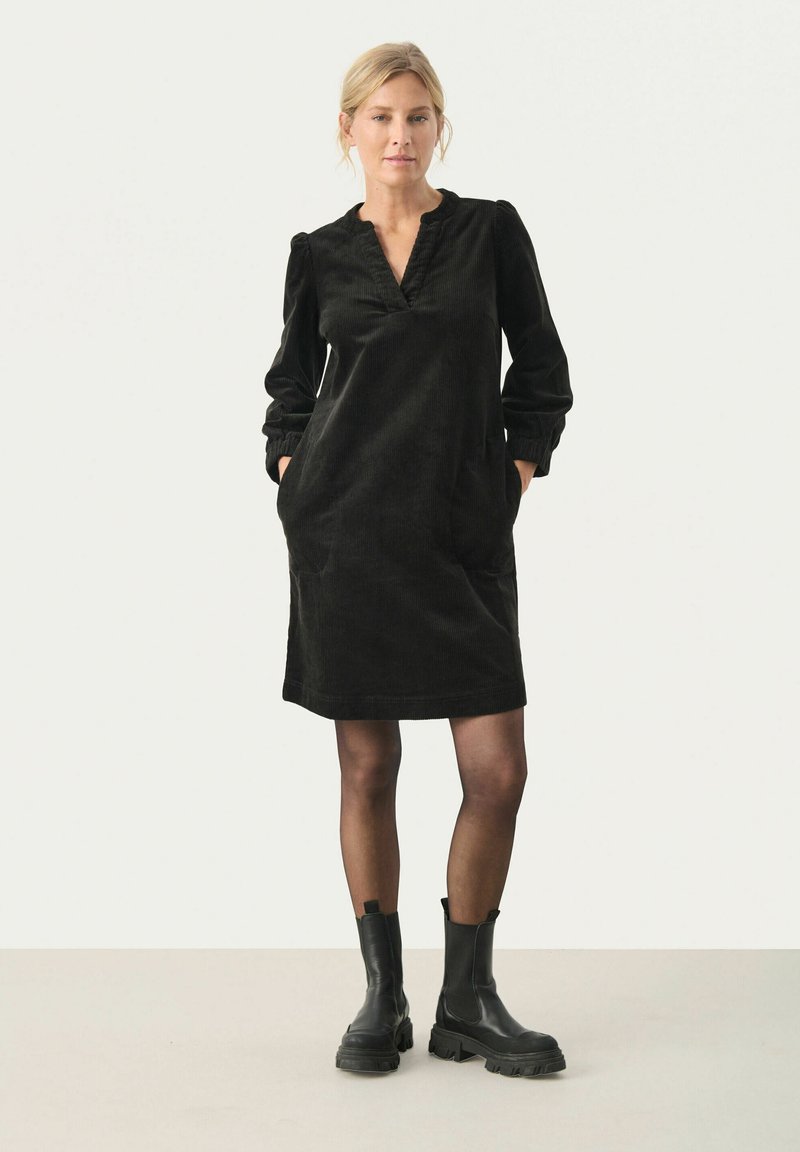 Robe en velours côtelé noire avec un décolleté en V, des manches bouffantes et des poches latérales. Associée à des bottines noires. Texture lisse et coupe décontractée.