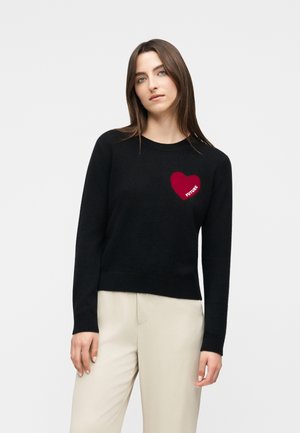 Femme portant un pull noir à manches longues avec un cœur rouge et le texte « FUTURE » sur la poitrine, vêtue d'un pantalon beige clair, debout devant un fond clair uni.