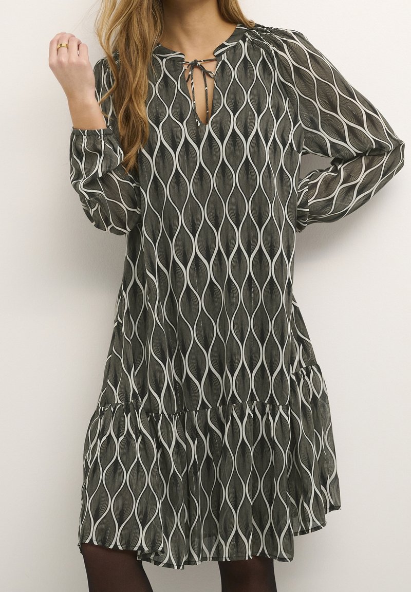 Robe ample à manches longues avec un motif vertical ondulé noir et blanc, un décolleté en forme de goutte noué avec un nœud, portée avec des collants foncés.