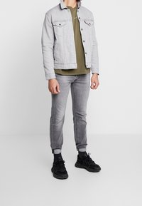 Grå denimjacka över en olivgrön skjorta, i kombination med grå jeans och svarta sneakers, med en avslappnad, figurnära design.