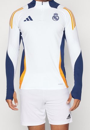 Mand iført en hvid og marineblå trøje med lange ærmer fra Real Madrid med orange accenter og hvide Adidas-shorts.