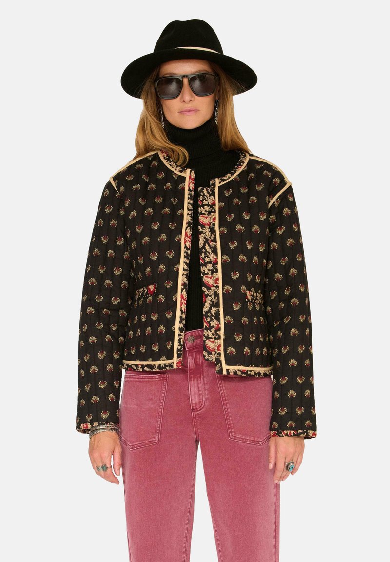 Veste matelassée noire avec broderies florales, bordure claire et col rond, portée avec un col roulé noir et un jean rose.