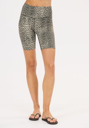 Mujer que lleva pantalones cortos de ciclismo de cintura alta con estampado de leopardo y chanclas negras, de pie frente a un fondo blanco liso.