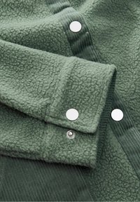 Gros plan de la manche d'une veste polaire verte avec des détails en tissu côtelé et des boutons-pression argentés au bas de la manche et à l'ouverture devant.