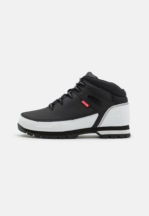 Bottes mi-hautes noir et blanc avec un matériau texturé, dotées de lacets, d'un accent rouge et d'une semelle robuste avec un logo en forme d'arbre.