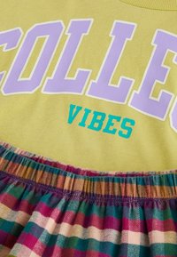 Gelbes Baumwolloberteil mit "COLLEGE" in Lila und "VIBES" in Türkis. Darunter ein mehrfarbiger karierter Stoff mit Pink-, Grün- und Brauntönen.