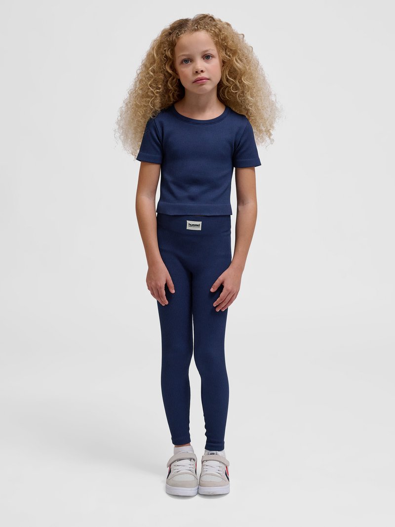 Młoda dziewczyna o kręconych włosach, ubrana w granatowy prążkowany krótki rękaw i legginsy, białe trampki, stojąca na jednolitym tle.