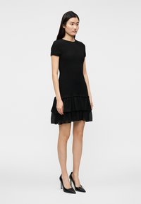 Μαύρο πλεκτό φόρεμα με κοντά μανίκια, εφαρμοστή κορυφή και στρωμένο τελείωμα με βολάν. Συνδυάζεται με μαύρες μυτερές γόβες.