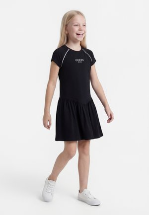 Fată zâmbitoare care merge, purtând o rochie neagră cu mâneci scurte cu logo-ul „GUESS” și adidași albi pe un fundal alb.