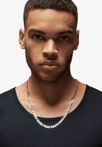 Zilveren ketting met schakels die in elkaar grijpen, gedragen over een zwart geribd tanktopje. Huidtint is medium met gezichtsbeharing en een neusring.