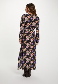 Maxi-jurk van zwarte stof met een bloemenpatroon in roze, paars en groen. Heeft lange mouwen en een getailleerde taille, gecombineerd met bruine laarzen.