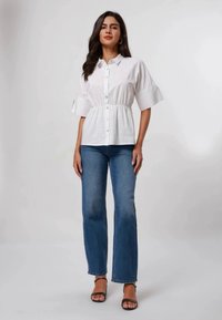Camicia bianca a maniche corte con bottoni e fascia elastica in vita, abbinata a jeans a zampa d'elefante blu e tacchi neri con cinturini.