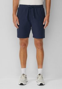 Marineblaue Shorts aus strukturiertem Stoff, mit elastischem Bund und Kordelzug. Tragen von hellen Sportschuhen und weißen Socken.