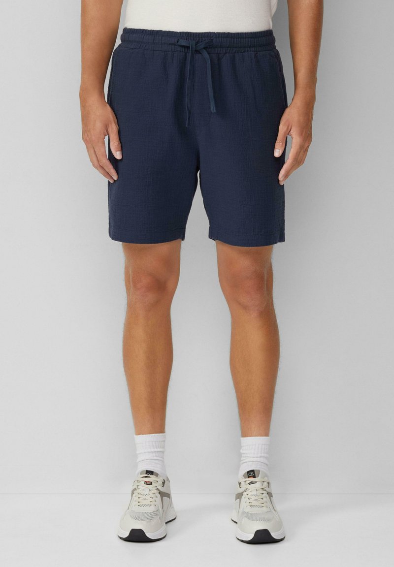 Marineblaue Shorts aus strukturiertem Stoff, mit elastischem Bund und Kordelzug. Tragen von hellen Sportschuhen und weißen Socken.