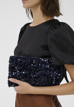 Navyblauer Clutch mit Pailletten, strukturiertem Oberflächen und Reißverschluss, in einer Hand gehalten. Glatte schwarze Bluse mit Puffärmeln sichtbar.