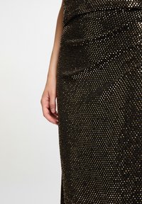 Un bras gauche et une main à côté d'un tissu de robe texturé noir et doré pailleté, coupé au niveau du torse, fond uni blanc cassé.
