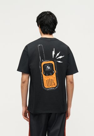 Persoon met donker haar die een zwart t-shirt draagt met een grote afbeelding van een oranje walkietalkie met signaallijnen op de achterkant.