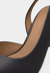 Chaussure à talon haut en cuir noir avec bride arrière, semelle intérieure beige et coutures visibles sur les bords.