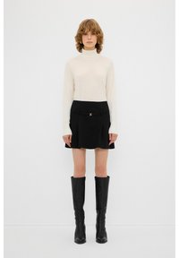Cremefarbener Rollkragensweater, schwarzer Faltenrock mit Schnallendetail, knielange schwarze Lederstiefel. Einfacher und körpernaher Stil.