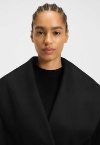 Black wool coat with oversized lapels, worn over a fitted black ribbed sweater. Smooth texture, minimal seams, and no visible patterns.