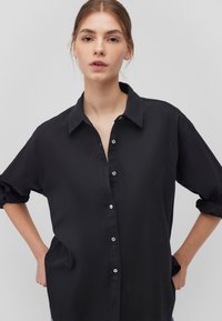 Black button-up shirt in lightweight fabric with a collared neck, rolled sleeves, and silver buttons, featuring a relaxed fit and side pockets.