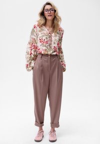 Bluse mit Blumenmuster und langen Ärmeln, V-Ausschnitt; beige hochgeschnittene Hose; rosa Turnschuhe; runde Brille.