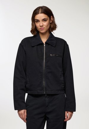 Giacca nera corta in tela, con colletto in corduroy, chiusura frontale con zip e una piccola tasca sul petto. Texture e design semplici.