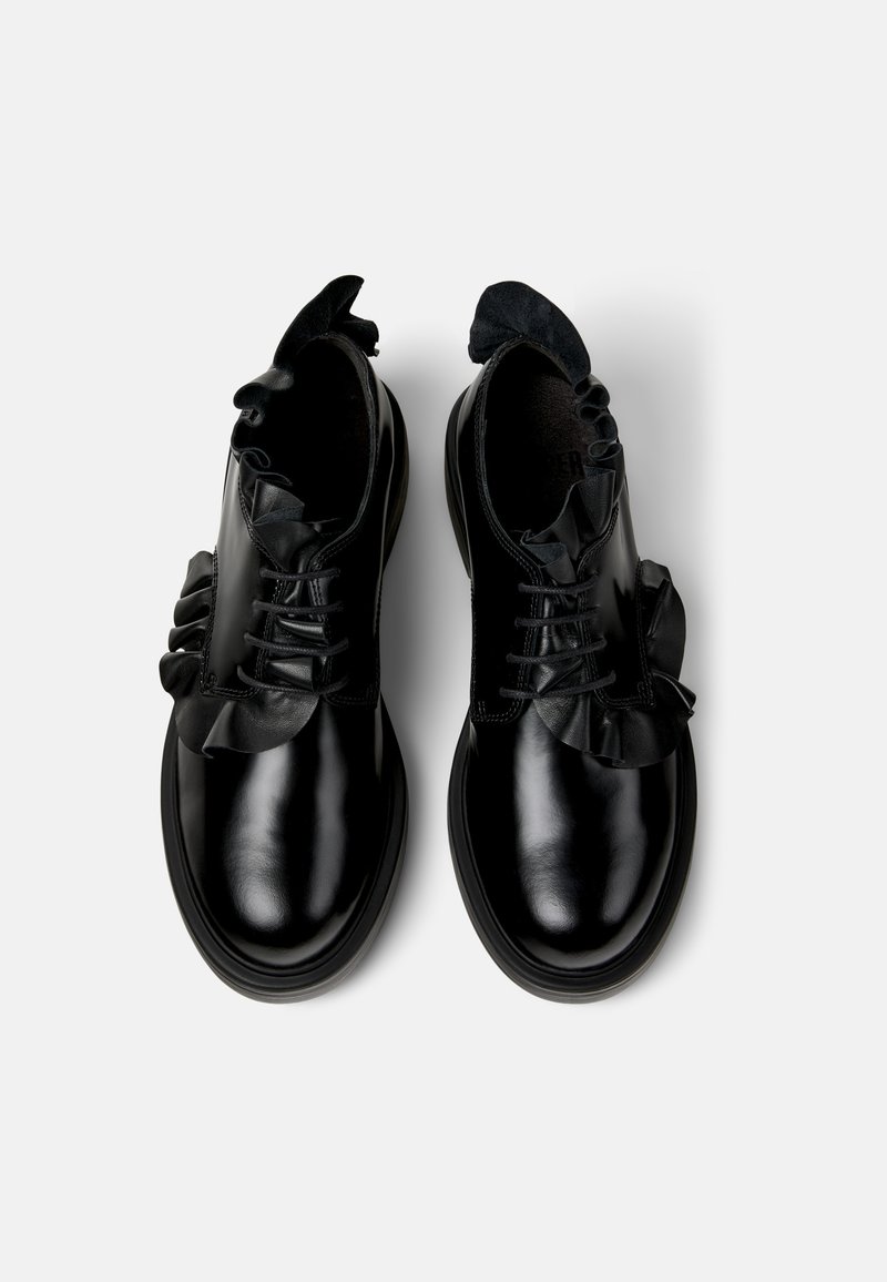 Sorte lædersko med en blank overflade, der har en rynket ruskindskant omkring snørebåndene og en rund tå. Flad gummisål.