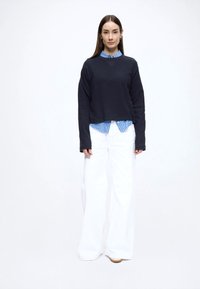 Tume sinine lühendatud kampsun, kihiliselt kantud hele sinise ja valge triibulise särgi peal, kombineeritud valgete laiade pükstega ja helepruunide kingadega.