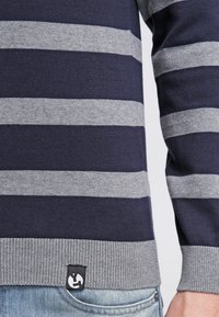 Maglione a righe blu navy e grigio realizzato in tessuto a maglia. Presenta un orlo a coste e un piccolo etichetta con logo in basso a sinistra.