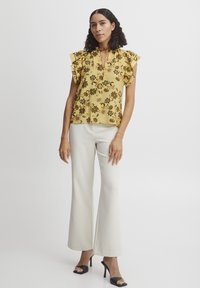 Blusa floreale gialla con maniche a sbuffo, scollatura a forma di chiave e vestibilità ampia, abbinata a pantaloni bianchi a gamba larga e sandali neri con tacco.