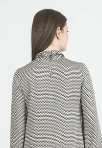 Halenka s dlouhým rukávem a vysokým límcem, s geometrickým vzorem v černé a krémové barvě. Zadní zip pro zapínání. Hladká textura látky.