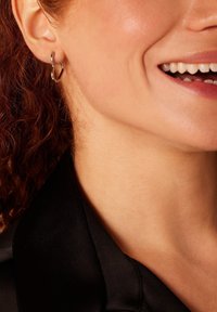 Zilveren oorring bevestigd aan een warm getinte huid, met een gladde en glanzende afwerking. Draagt een zwarte satijnen kraag.